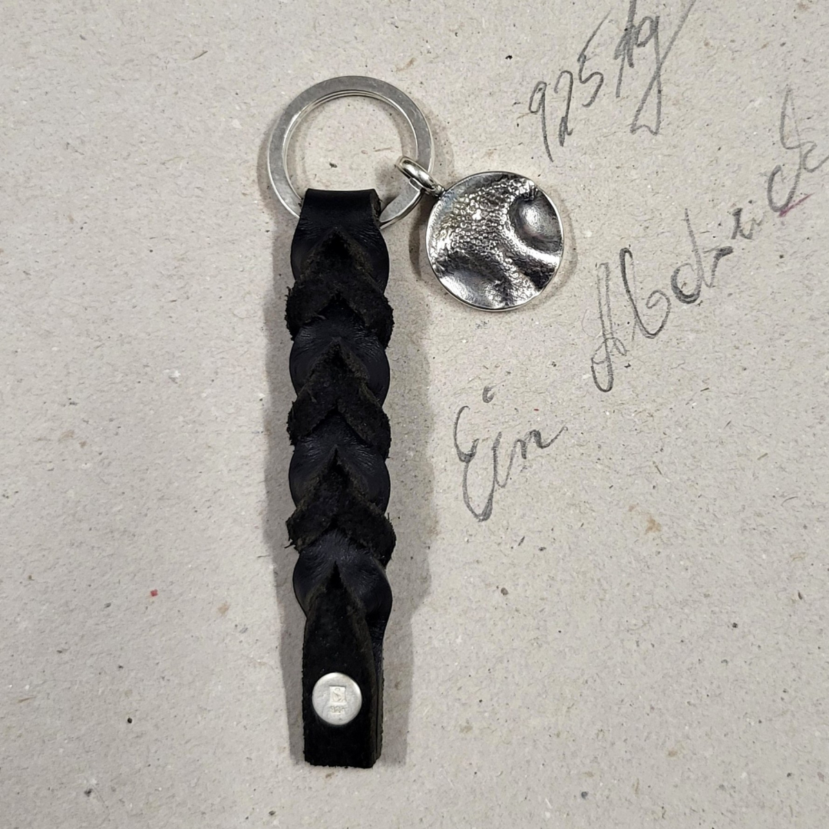 Handgefertigter runder Hundenasen-Anhänger aus 925er Silber an einem Schlüsselring mit einem schwarzen, geflochtenen Lederband auf einer hellen Unterlage mit handschriftlicher Notiz.
