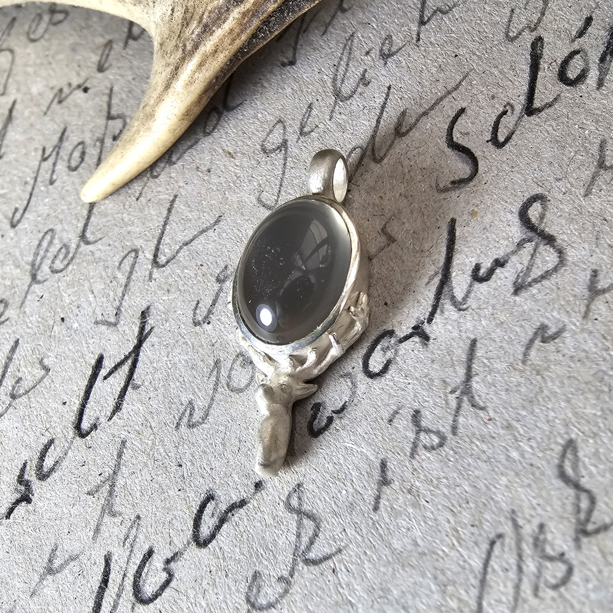 Hirschkopf-Anhänger aus 925 Silber mit grauem indischem Mondstein auf grauer Pappe mit jagdlichem Bleistifttext