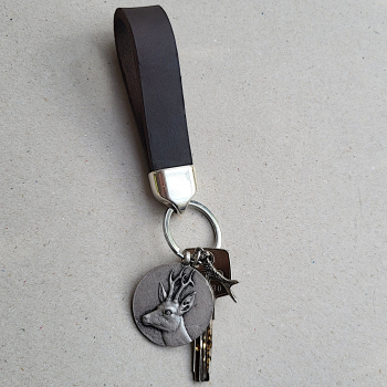 Handgefertigter Schlüsselanhänger aus braunem Lederband mit rundem Rehbock-Anhänger aus Metall, liegend auf hellem Untergrund.