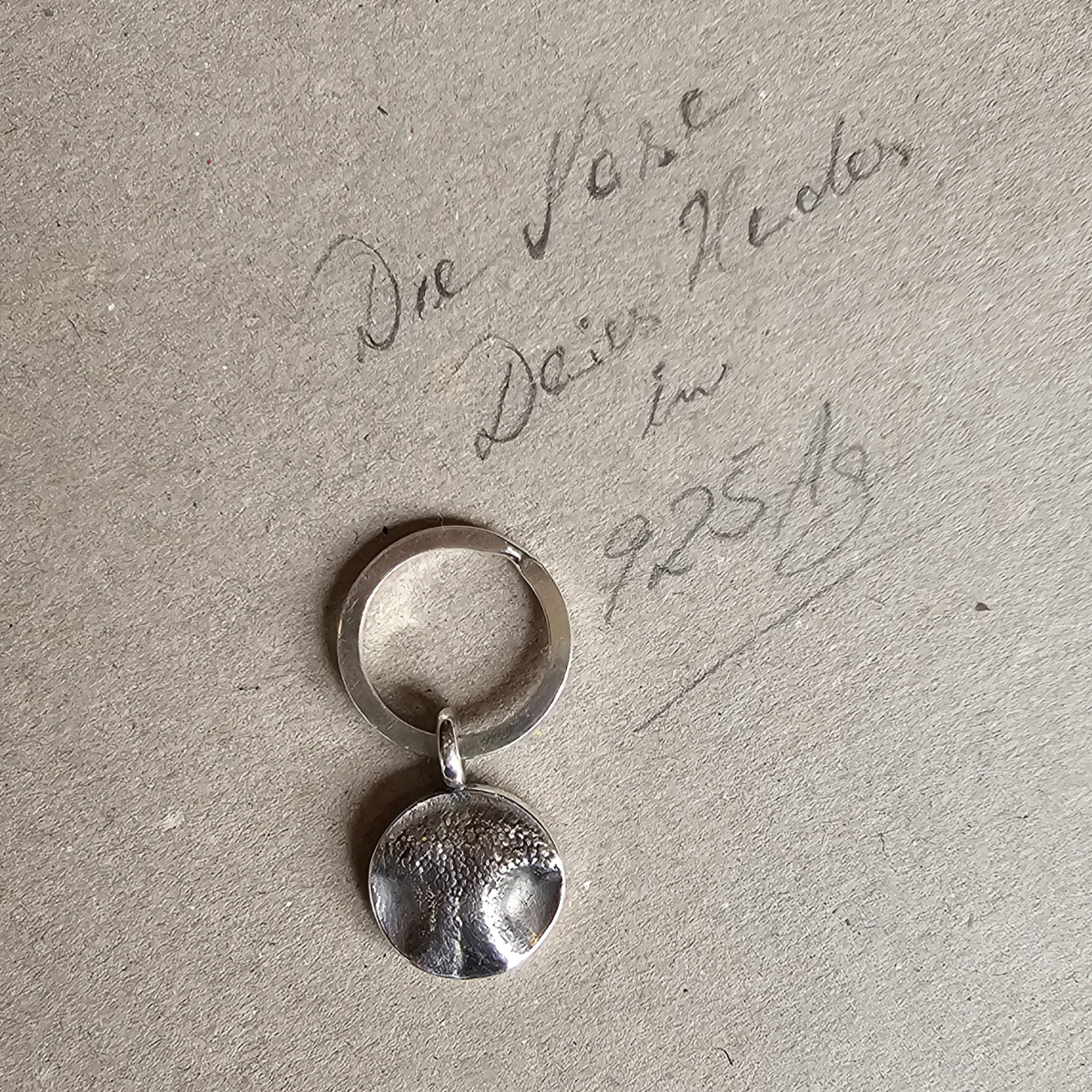 Ein handgefertigter runder Hundenasen-Anhänger aus echtem Silber an einem Schlüsselring, abgelegt auf einer hellbraunen Pappunterlage mit der handschriftlichen Notiz Die Nase Deines Hundes in 925 Ag.